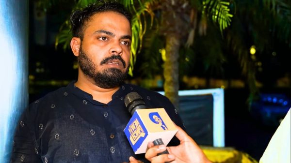 Man with a beard in a dark kurta speaks into a microphone during an outdoor interview at night.