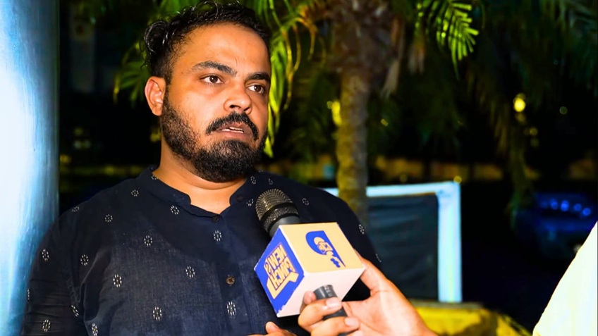 Man with a beard in a dark kurta speaks into a microphone during an outdoor interview at night.