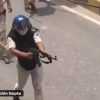 Person wearing a helmet and protective vest aims a rifle on a street, with another person nearby in the background.