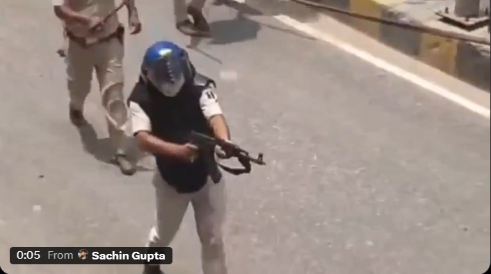 Person wearing a helmet and protective vest aims a rifle on a street, with another person nearby in the background.
