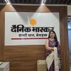 Smiling woman in a purple saree standing beside the Dainik Bhaskar sign on a wooden wall in an office.