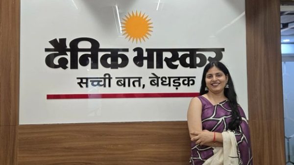 Smiling woman in a purple saree standing beside the Dainik Bhaskar sign on a wooden wall in an office.