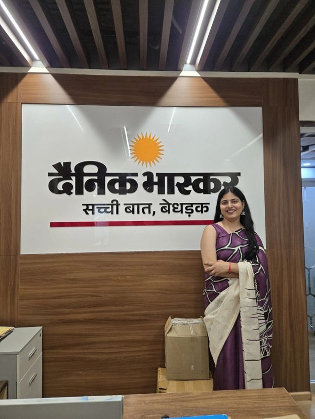 Smiling woman in a purple saree standing beside the Dainik Bhaskar sign on a wooden wall in an office.