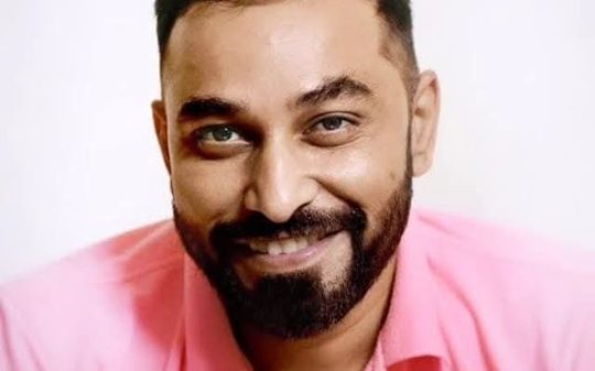 Smiling man with short dark hair and beard wearing a pink shirt against a light background.