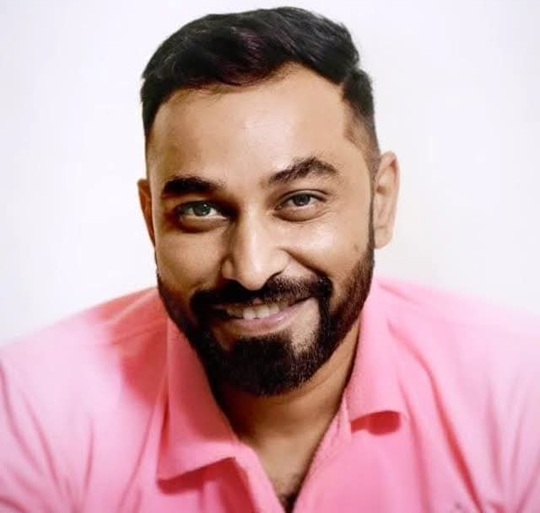 Smiling man with short dark hair and beard wearing a pink shirt against a light background.