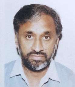 Head-and-shoulders portrait of a middle-aged man with gray beard, wearing a light blue shirt, looking at the camera.