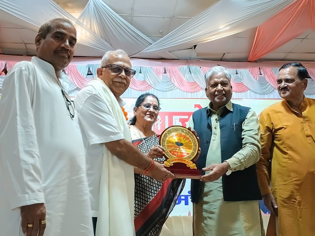 Group of five people on a stage presenting a circular plaque award to a man in a green shirt and vest, smiles all around.