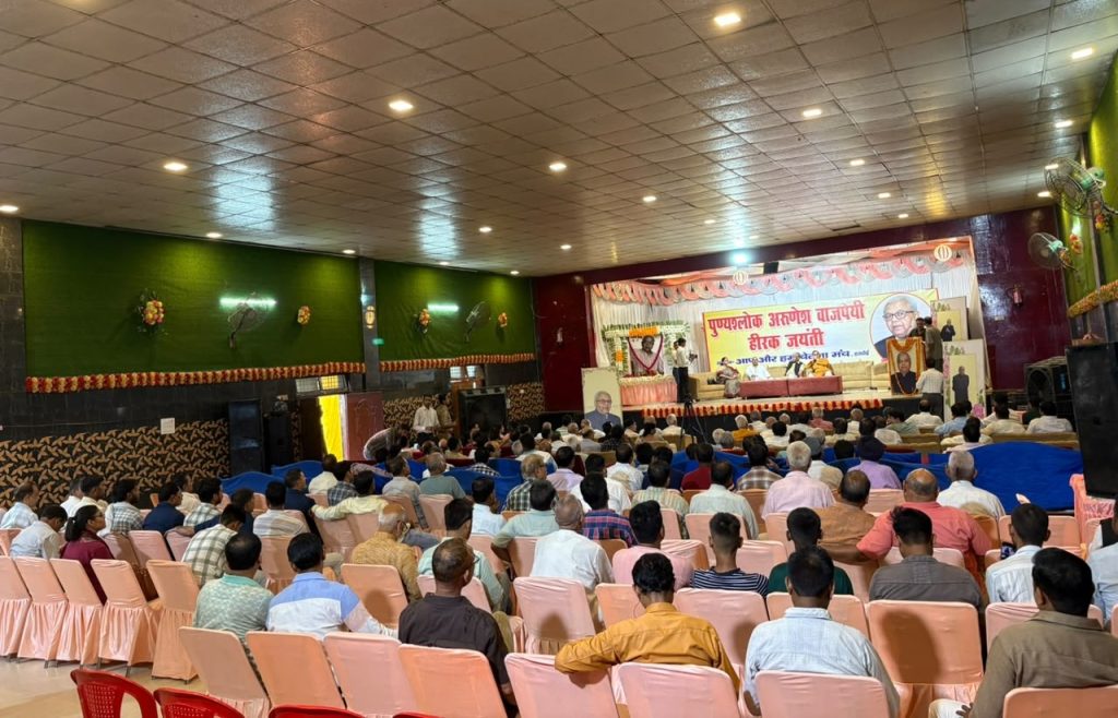 Audience seated in a decorated hall watching a stage program, with a large Hindi banner and portraits on the backdrop.