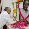 Man in a white shirt places pink flower petals on a table in front of a large portrait of an elderly man adorned with garlands, during a memorial tribute.