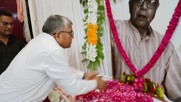 Man in a white shirt places pink flower petals on a table in front of a large portrait of an elderly man adorned with garlands, during a memorial tribute.