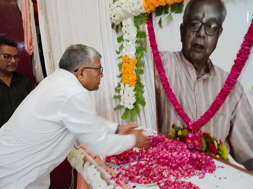 Man in a white shirt places pink flower petals on a table in front of a large portrait of an elderly man adorned with garlands, during a memorial tribute.