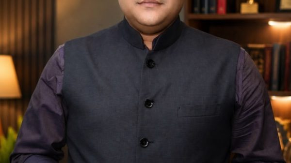 Portrait of a man in a dark vest and purple shirt, standing in a study with bookshelves behind him.