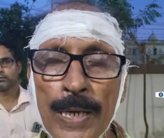 Man with a white head bandage and black glasses speaking to the camera outdoors, with a blurred man in the background.