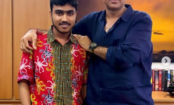 Two men stand arm in arm indoors, the younger in a bright red patterned shirt and white shorts, the older in a dark blue shirt and light pants, smiling at the camera.