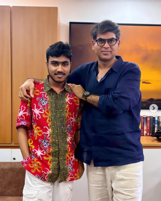 Two men stand arm in arm indoors, the younger in a bright red patterned shirt and white shorts, the older in a dark blue shirt and light pants, smiling at the camera.