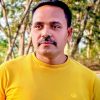 Portrait of a man with a mustache wearing a yellow shirt, outdoors with trees in the background, looking slightly to the side.