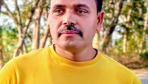 Portrait of a man with a mustache wearing a yellow shirt, outdoors with trees in the background, looking slightly to the side.