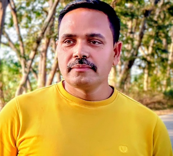 Portrait of a man with a mustache wearing a yellow shirt, outdoors with trees in the background, looking slightly to the side.