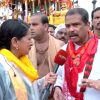 Female journalist interviews a man in a white shirt with a red scarf at a festival; NDTV microphone, crowd and decorative floats in the background.
