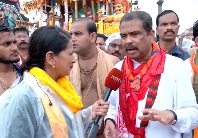 Female journalist interviews a man in a white shirt with a red scarf at a festival; NDTV microphone, crowd and decorative floats in the background.