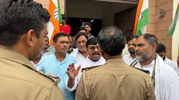 Group of men gathered outside a building, Indian flags raised, police in tan uniforms in the foreground.