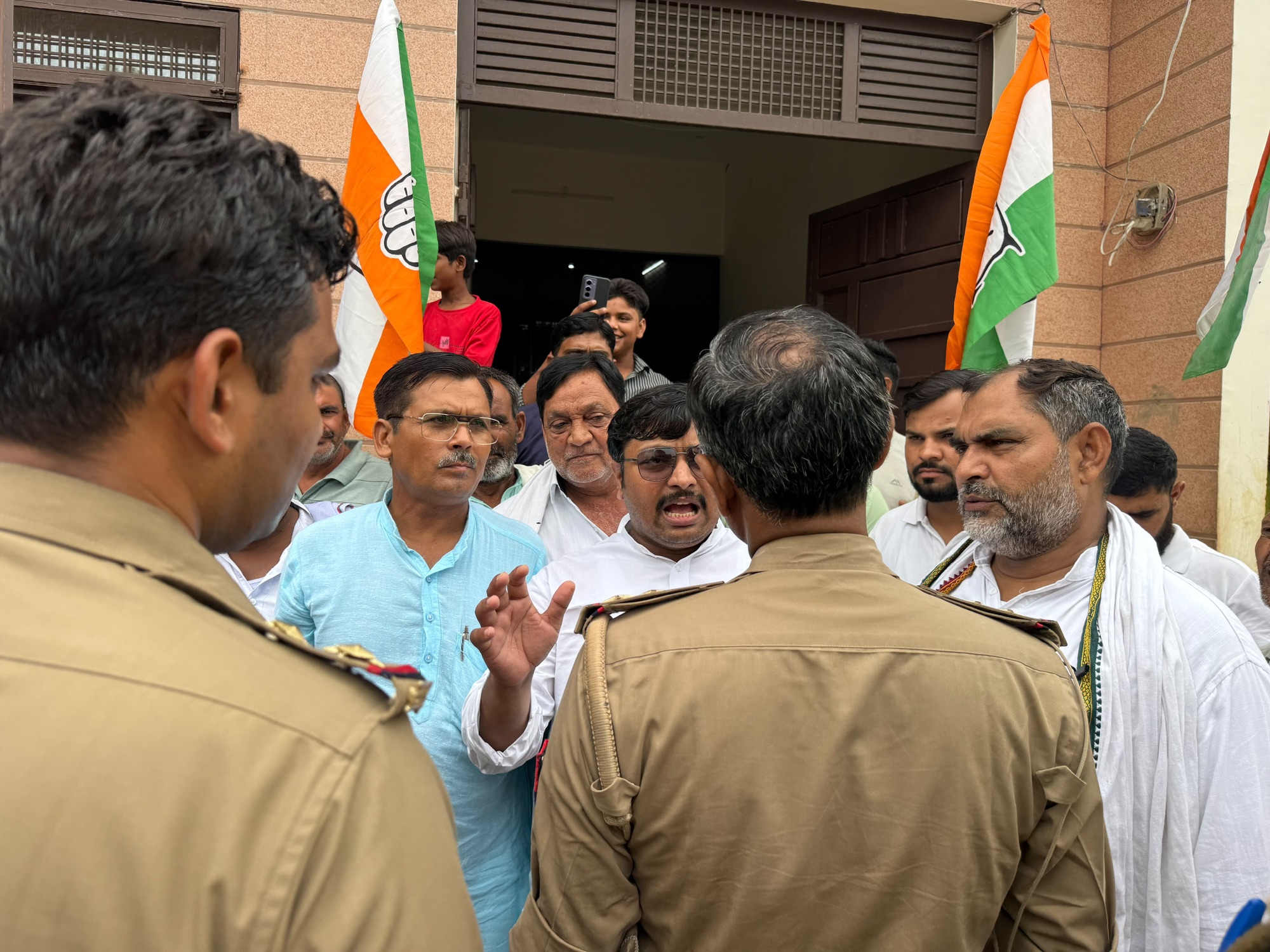 Group of men gathered outside a building, Indian flags raised, police in tan uniforms in the foreground.