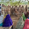 Police officers stand in a line addressing a group of seated women in colorful sarees under a tree in a rural outdoor area.