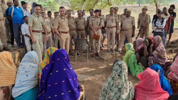 Police officers stand in a line addressing a group of seated women in colorful sarees under a tree in a rural outdoor area.