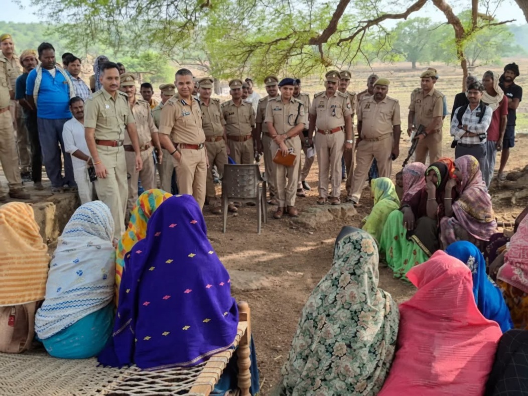 Police officers stand in a line addressing a group of seated women in colorful sarees under a tree in a rural outdoor area.