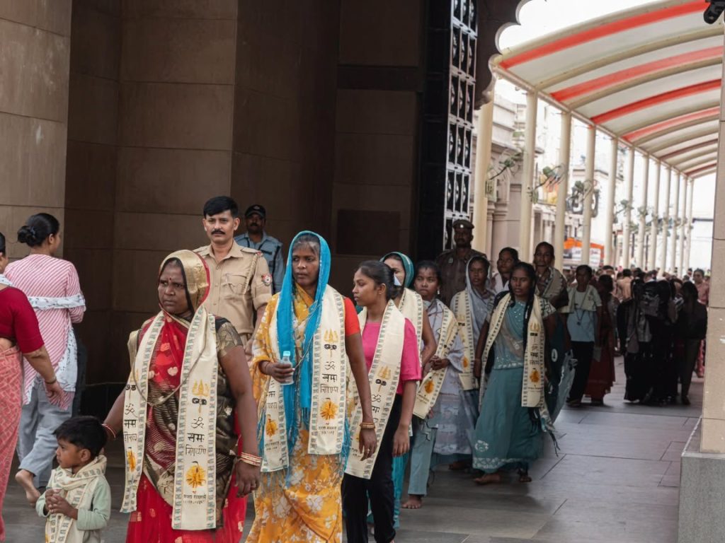 chandauli_2 | No. 1 Indian Media News Portal Line of Indian women and children in colorful sarees with ceremonial scarves, walking in a procession under a covered walkway, escorted by police.