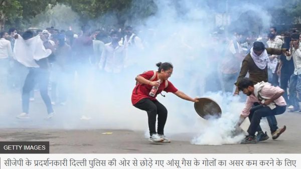 Young woman in a red shirt throws or hands a bottle toward a protester with a shield amid tear gas at a street demonstration.