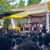 Large crowd at an outdoor stage as speakers address a rally, with a small Indian flag in front and colorful tarps overhead.