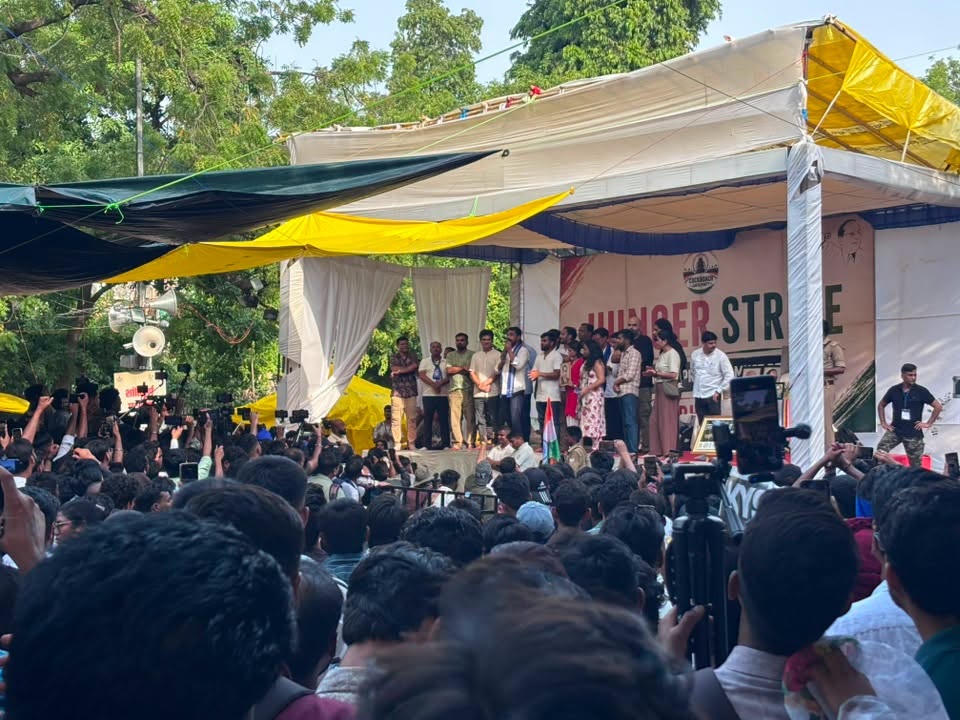 Large crowd at an outdoor stage as speakers address a rally, with a small Indian flag in front and colorful tarps overhead.