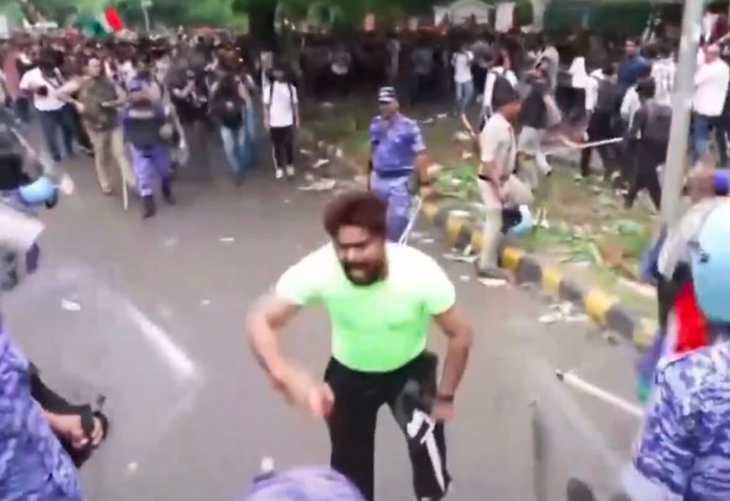 Screenshot | No. 1 Indian Media News Portal Man in neon green shirt moves toward the camera on a crowded street; bystanders and police in the background.