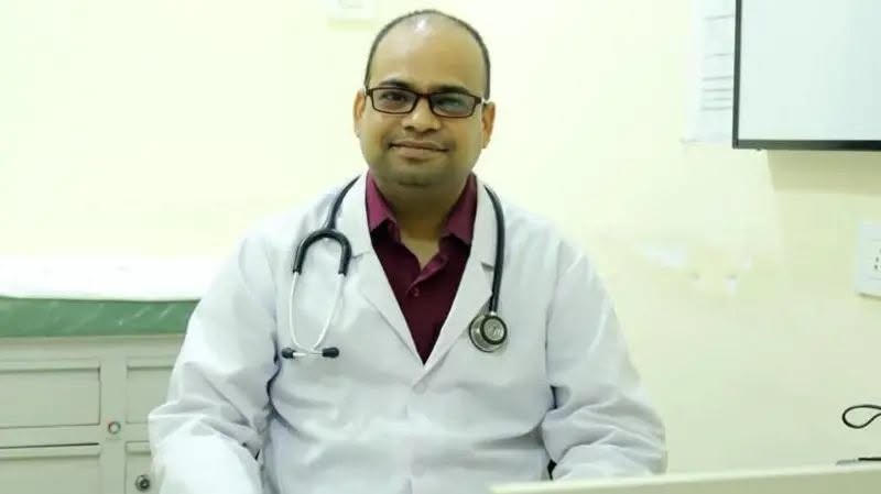 debta | No. 1 Indian Media News Portal Male doctor in a white coat with a stethoscope around his neck, sitting at a desk in a clinic and smiling at the camera.