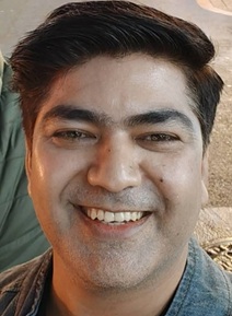 Close-up of a man with dark hair smiling outdoors, wearing a blue denim jacket.