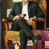 Man in a black suit sits on an ornate wooden chair on stage, holding a red microphone during a panel or interview event with banners in the background.