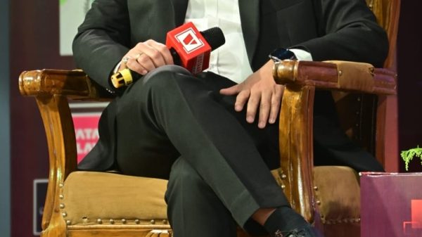 Man in a black suit sits on an ornate wooden chair on stage, holding a red microphone during a panel or interview event with banners in the background.