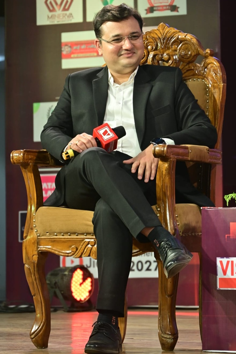 Man in a black suit sits on an ornate wooden chair on stage, holding a red microphone during a panel or interview event with banners in the background.