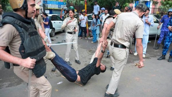 Police officers in beige uniforms and protective gear restrain a man being dragged by his arms while onlookers watch on a city street