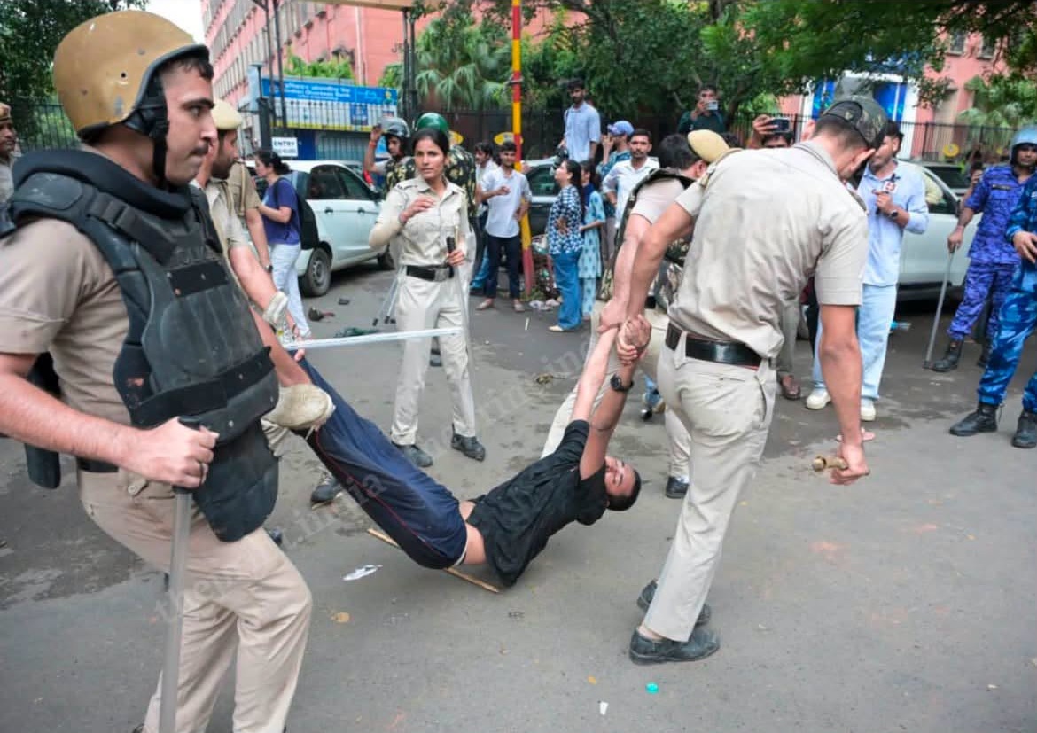 Police officers in beige uniforms and protective gear restrain a man being dragged by his arms while onlookers watch on a city street