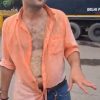 Man in an orange unbuttoned shirt exposing his chest, gesturing with his right hand on a city street; a large truck and trees behind him, and keys hanging from his belt loop.