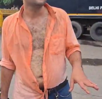 Man in an orange unbuttoned shirt exposing his chest, gesturing with his right hand on a city street; a large truck and trees behind him, and keys hanging from his belt loop.