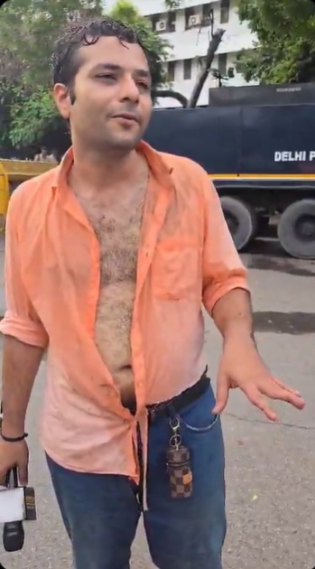 Man in an orange unbuttoned shirt exposing his chest, gesturing with his right hand on a city street; a large truck and trees behind him, and keys hanging from his belt loop.