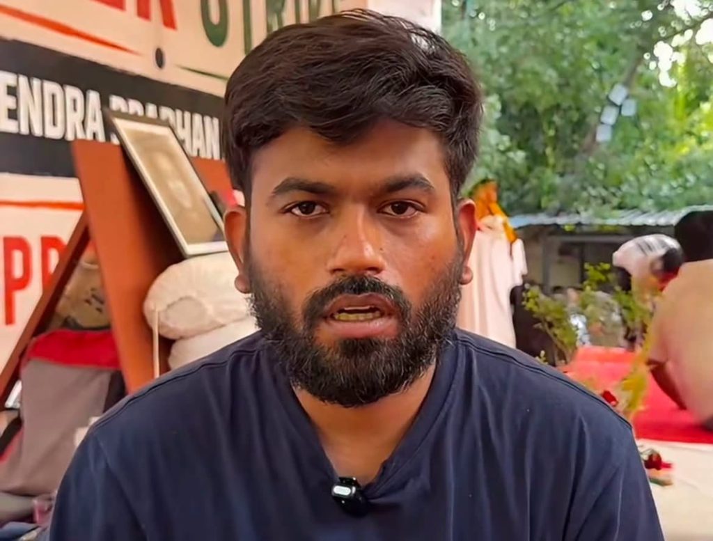Screenshot | No. 1 Indian Media News Portal Close-up of a bearded man in a dark blue shirt speaking to camera at an outdoor event with greenery in the background