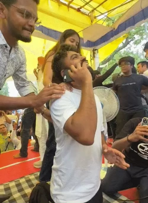 A man in a white t-shirt is on his phone while being surrounded by people under a yellow canopy at an outdoor event.