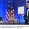At a formal event, a man in a tuxedo presents a sign reading 'Jerry Garcia' while a woman in a sparkling gown sits at a table with wine glasses; an American flag and blue backdrop are behind them.