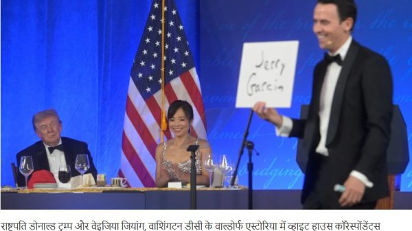 At a formal event, a man in a tuxedo presents a sign reading 'Jerry Garcia' while a woman in a sparkling gown sits at a table with wine glasses; an American flag and blue backdrop are behind them.