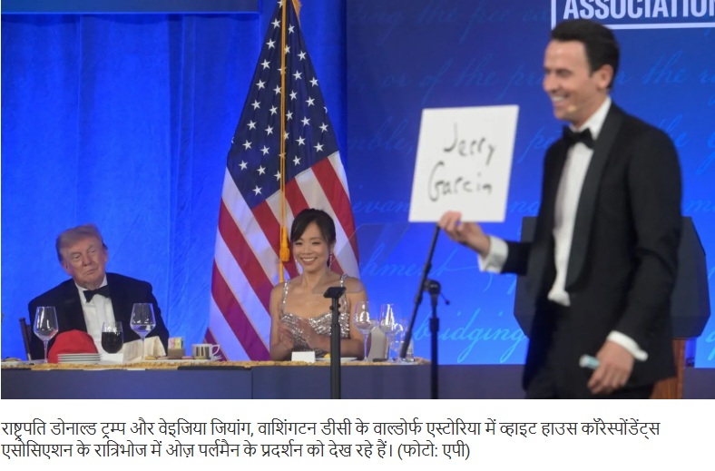 At a formal event, a man in a tuxedo presents a sign reading 'Jerry Garcia' while a woman in a sparkling gown sits at a table with wine glasses; an American flag and blue backdrop are behind them.
