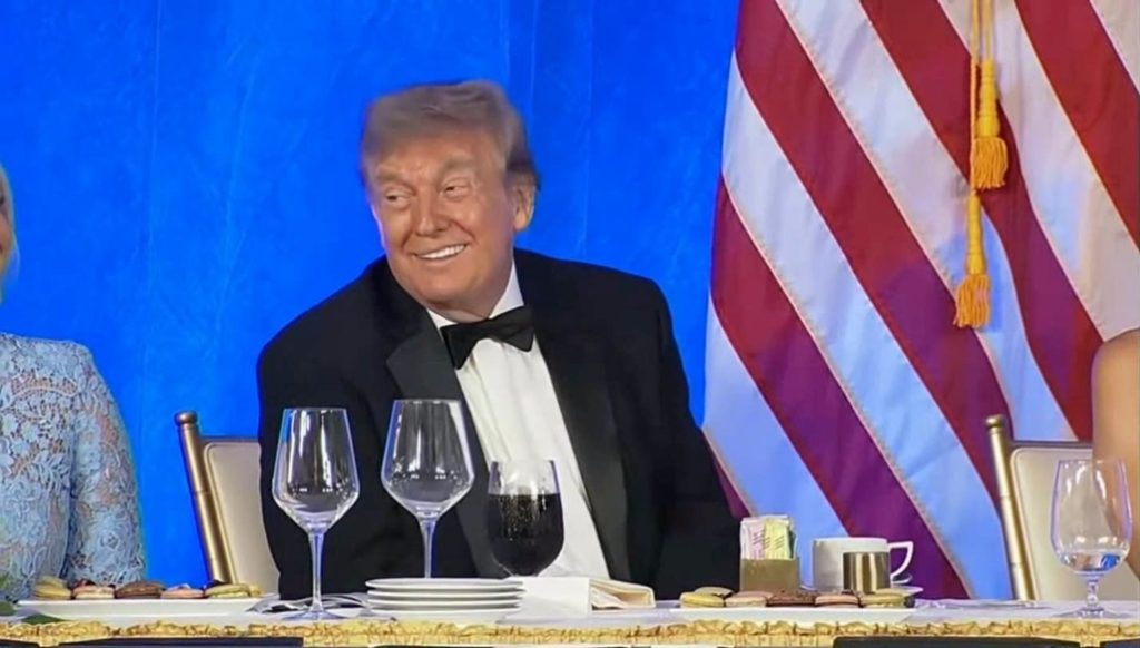 donald_1 | No. 1 Indian Media News Portal Man in a black tuxedo and bow tie smiles at a formal dinner, with two wine glasses and an American flag in the blue backdrop behind him.